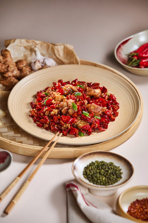 A plate of Sichuanese spicy wok-fried chicken with chilli. Garlic bulbs, ginger and whole fresh chillis are placed around the dish.