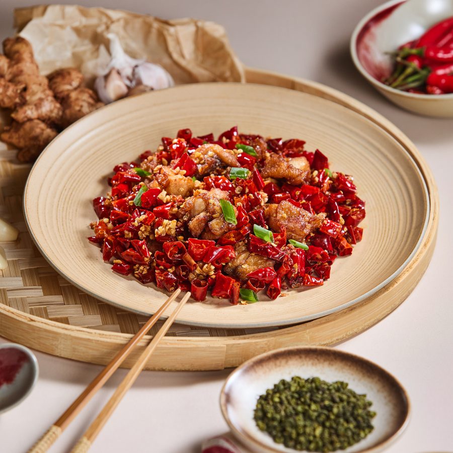A plate of Sichuanese spicy wok-fried chicken with chilli. Garlic bulbs, ginger and whole fresh chillis are placed around the dish.