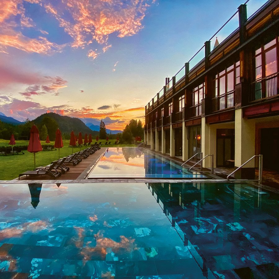 Exterior view at sunset of an outdoor pool, with grass and red umbrellas in the background. 