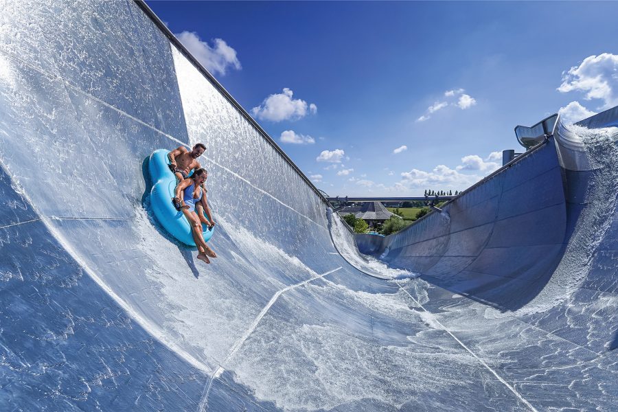 Exterior view of a couple riding a rubber ring down a large, blue water slide during the day.
