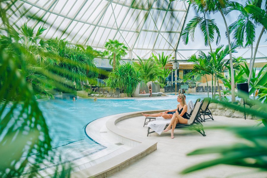 Interior view of a woman in a black swimsuit reclining on a deck chair beside a tropical spa pool, surrounded by lush green plants.