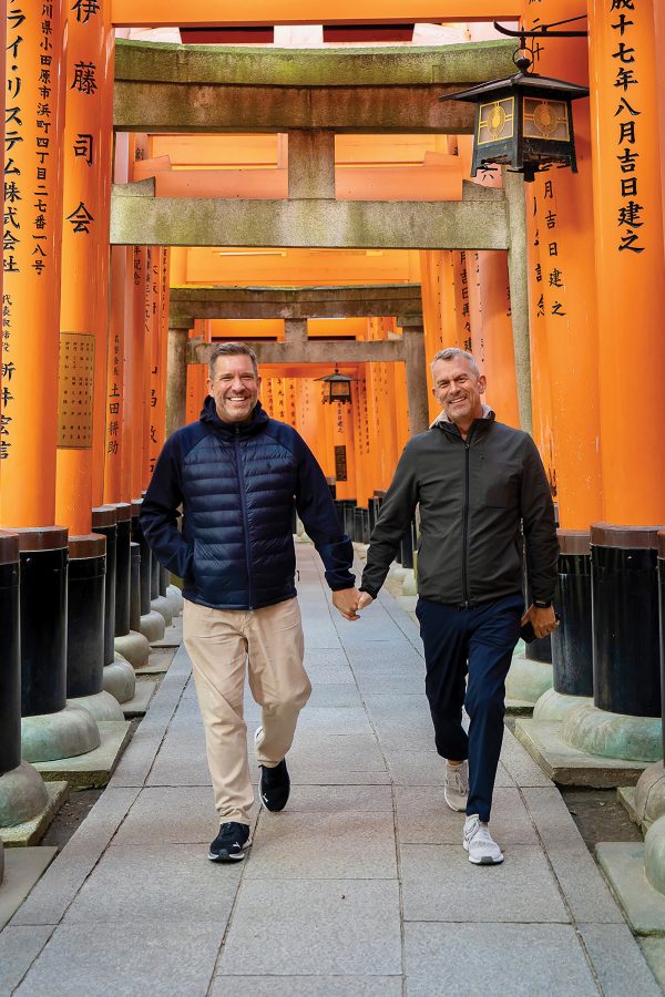 Two men hold hands and smile as they walk through Japanese religious torii gates.