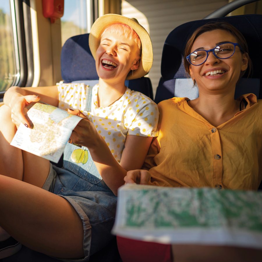 Two women laugh as they sit side by side on a train with maps in their hands