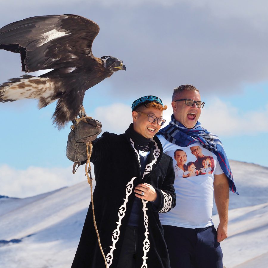 Two men smile as one holds out his arm with a falcon on it, against a snowy mountain backdrop.