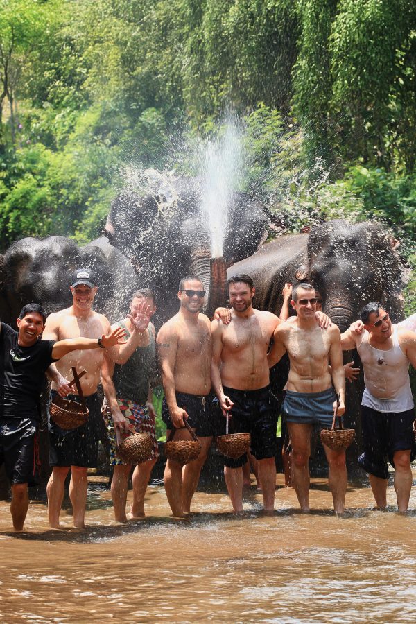 A group of men in swimming trunks smile as they pose in a river with water bursting overhead behind them.