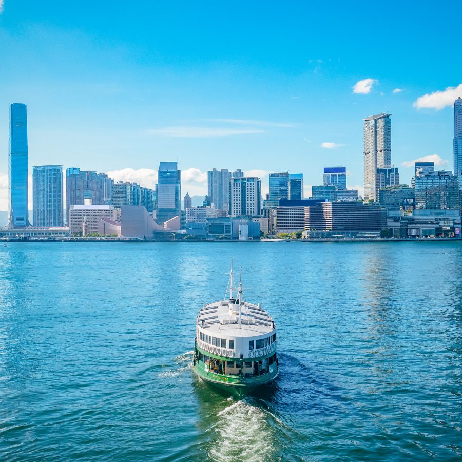 The Star Ferry carves through the water of Victoria Harbour as skyscrapers on Kowloon stand tall on the horizon. 