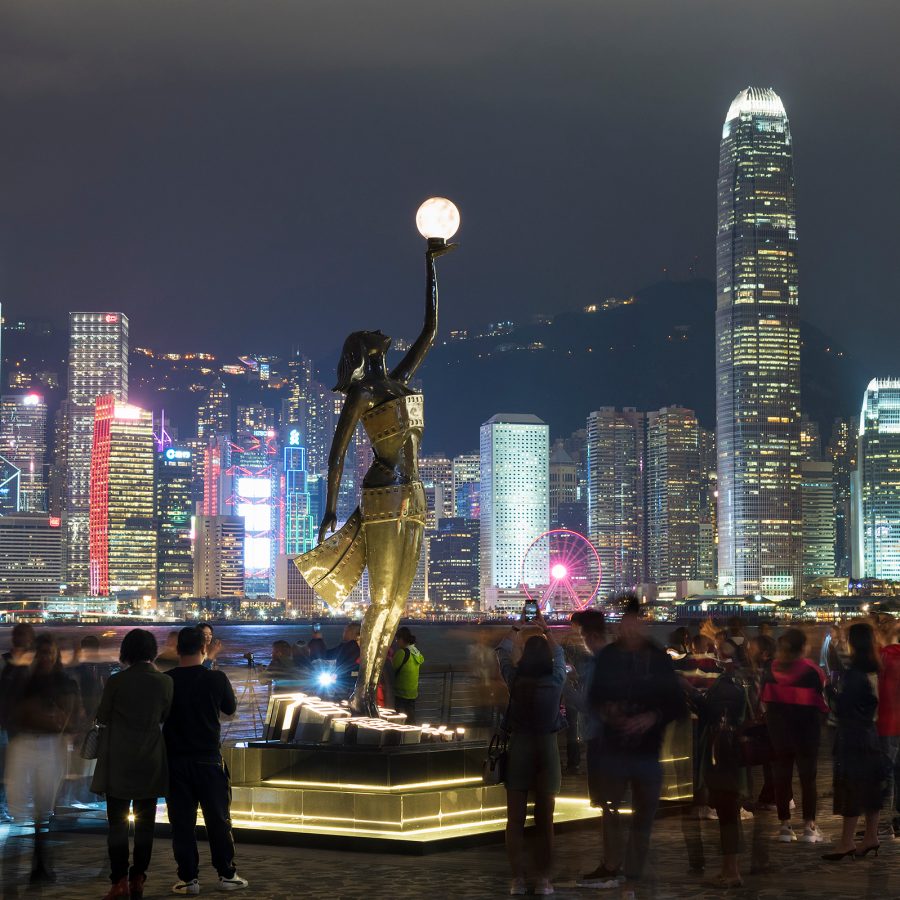 Visitors to the Avenue of Stars admire a statue at night as the skyline of Hong Kong Island glitters across the harbour.