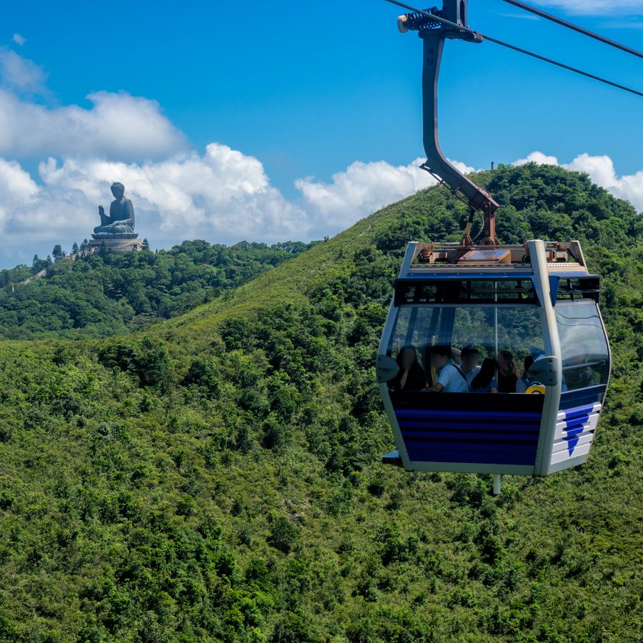 A Ngong Ping 360 cable car is suspended in the air against the backdrop of a lush green mountain, the Tian Tan Buddha visible in the distance. 