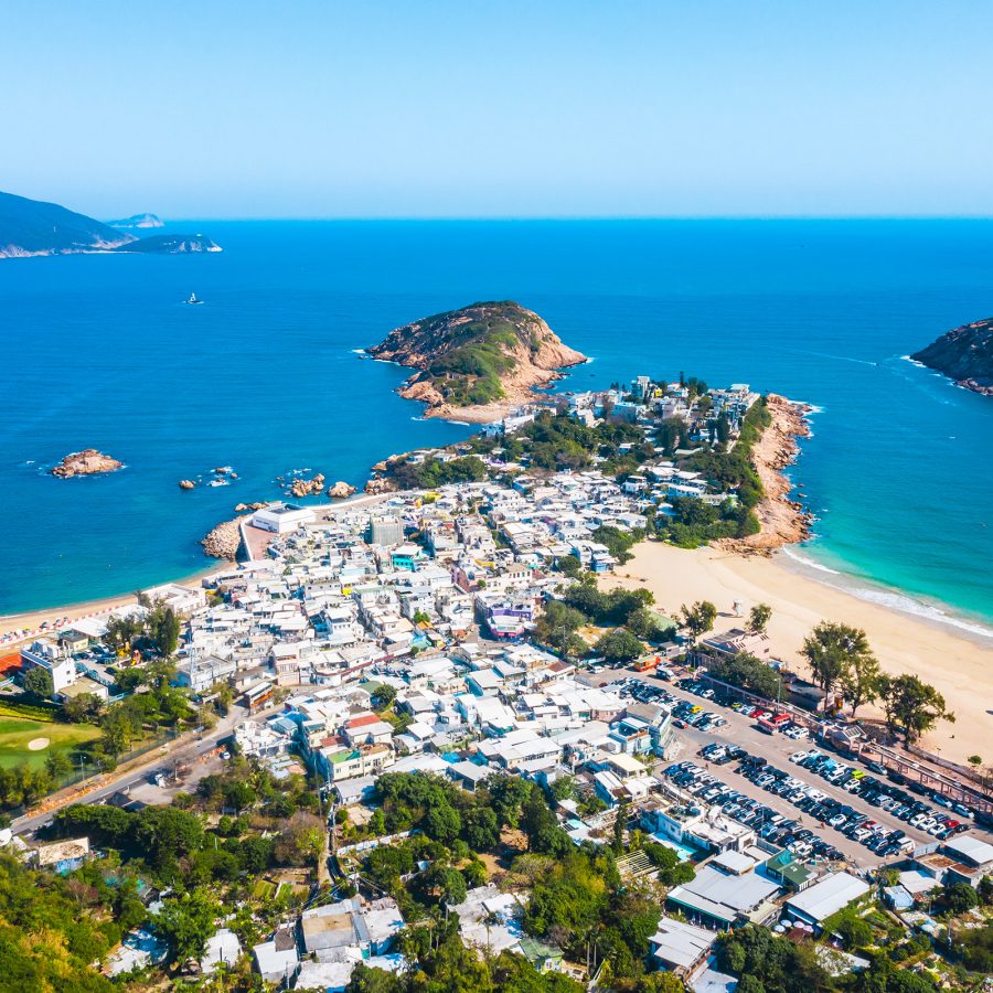 A bird’s eye view of Shek O and Shek O Beach shows bunched up low white buildings surrounded by golden beaches giving way to the surrounding sea. 