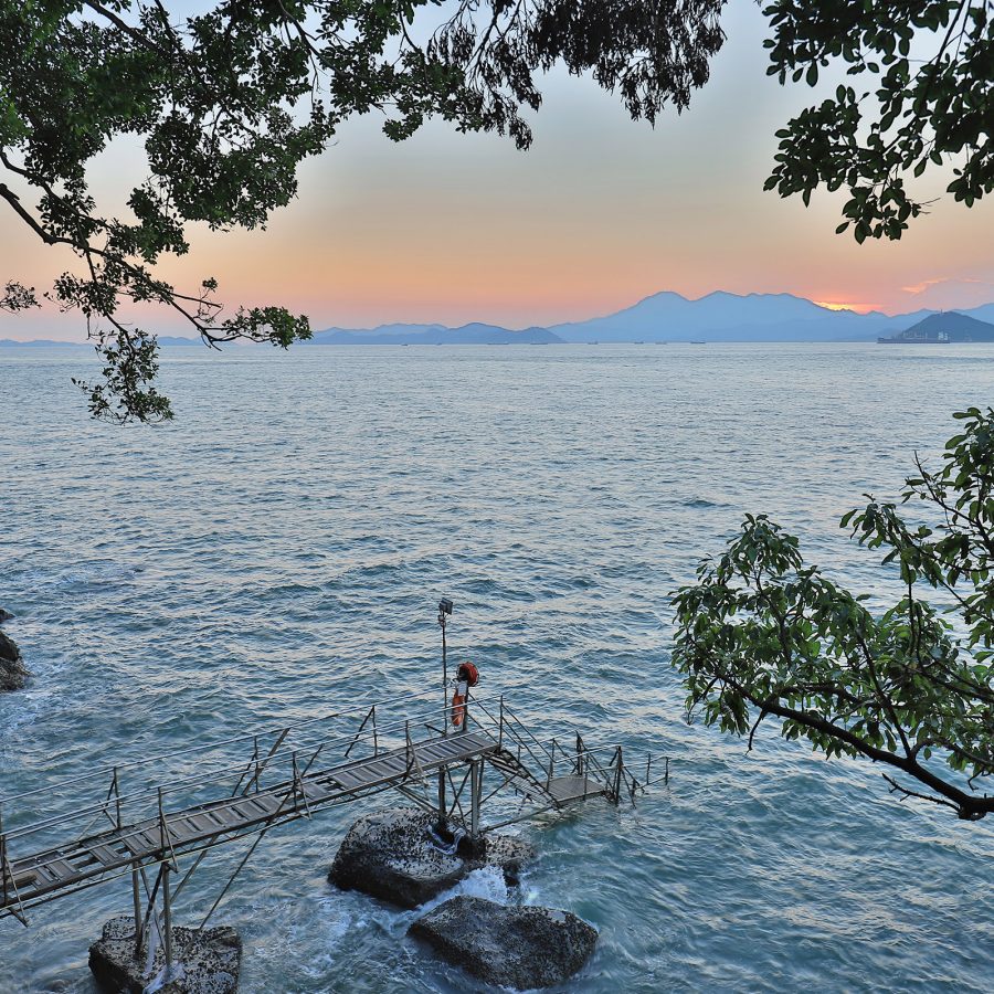 A view from Sai Wan Swimming Shed shows the sun setting behind distant mountains across the water.  
