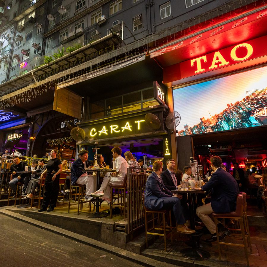 Signs of bars huddled next to each other shine enticingly as diners sit at outdoor tables. 