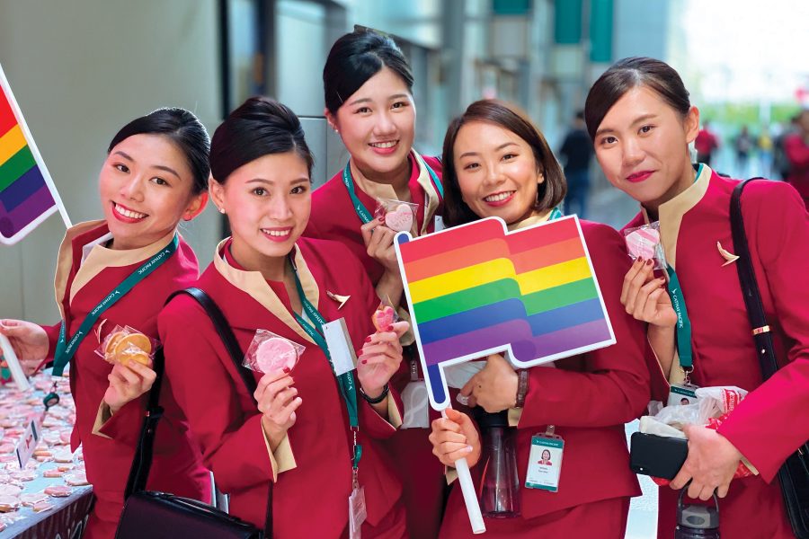 Five female flight attendants hold up signs of rainbow Pride flags as they smile for the camera.