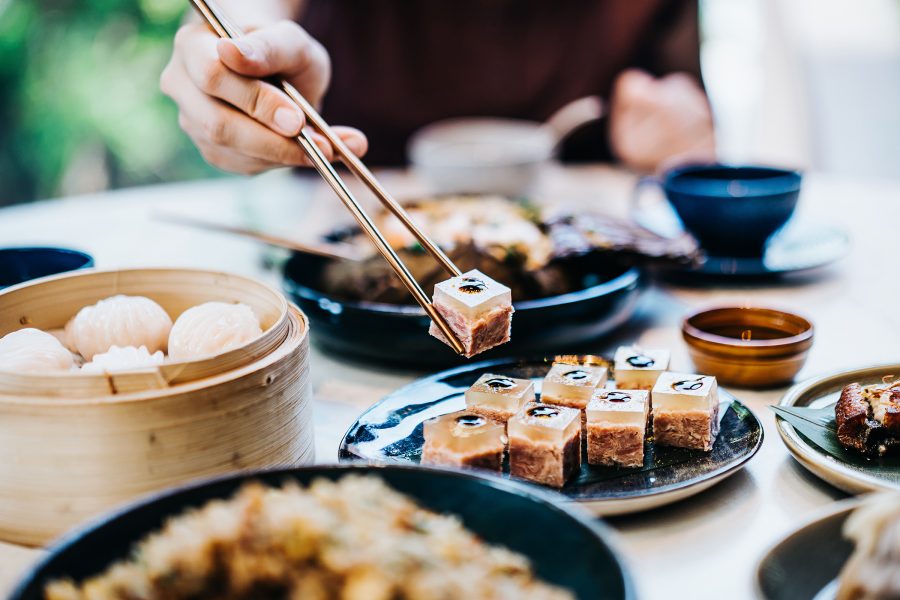 A person picks up a piece of pork with chopsticks. The table is laden with a bamboo steamer of har gau and other dishes.