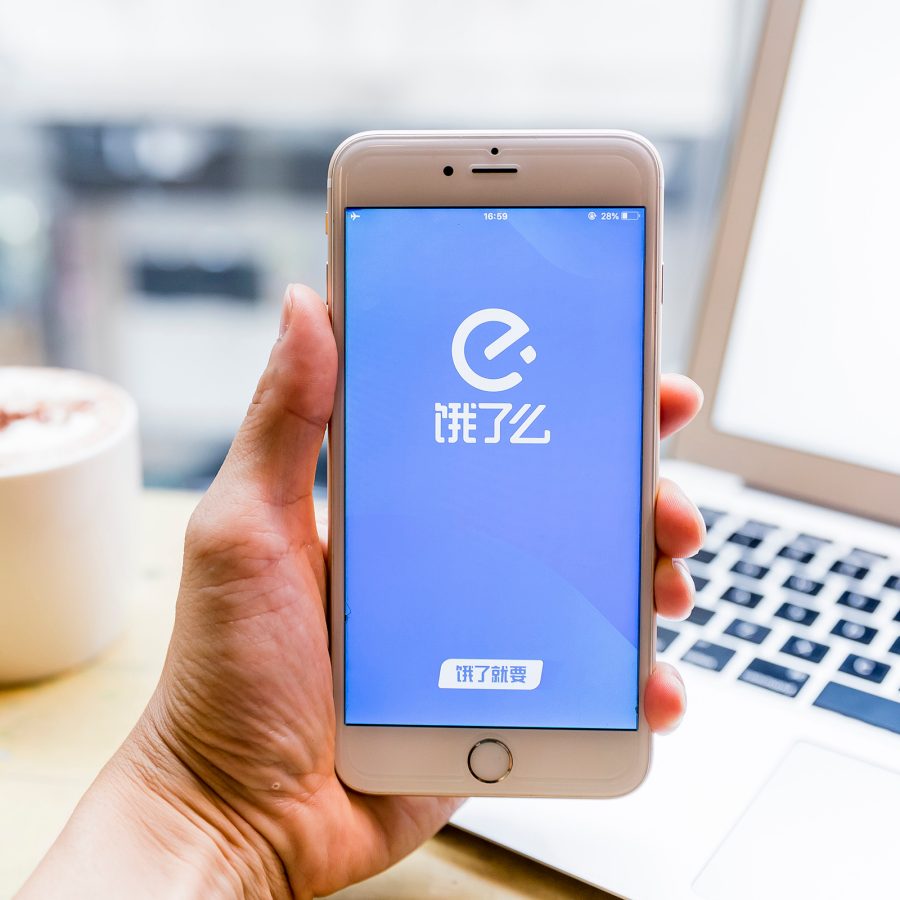 A hand holding a smartphone with the Ele.me app open. A laptop and cup of coffee are in the background.