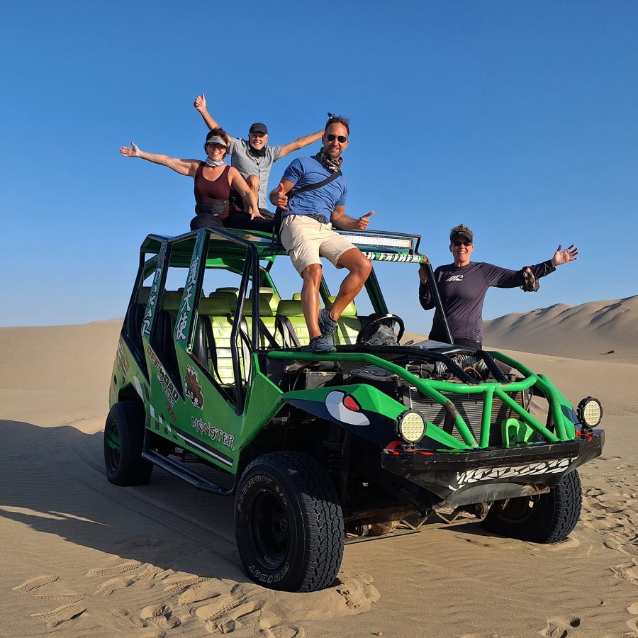 A small group of LGBT+ travellers sit on top of a car in the desert with their arms raised