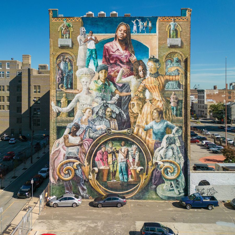 Exterior view of a large, colourful mural on the side of a building, with blue sky and cars parked below. The mural is titled Common Threads and it depicts women from different centuries wearing contemporary clothes and period clothes