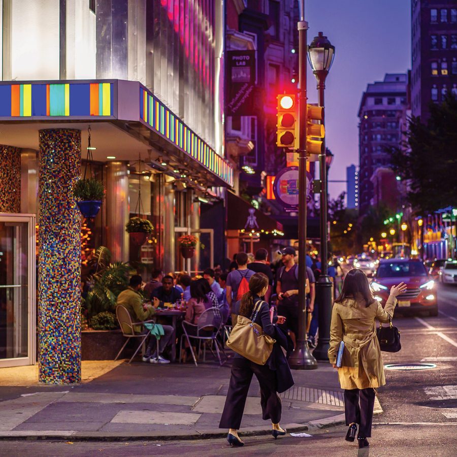 Exterior view of two people crossing the street in Midtown Village at dusk as other people sit outside a restaurant