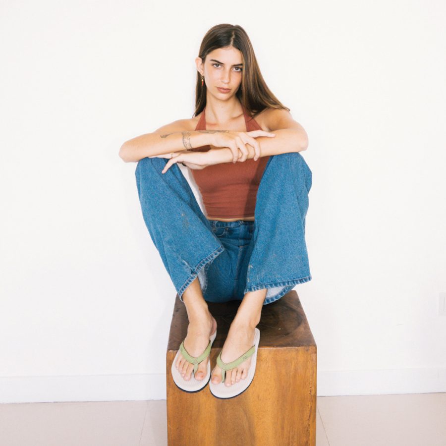 A model crossing her arms on a wooden seat shows off a pair of sandals from Indosole.