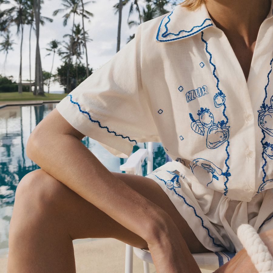 A person sitting outside next to a pool sports a white button up embroidered with blue line art from Magali Pascal.