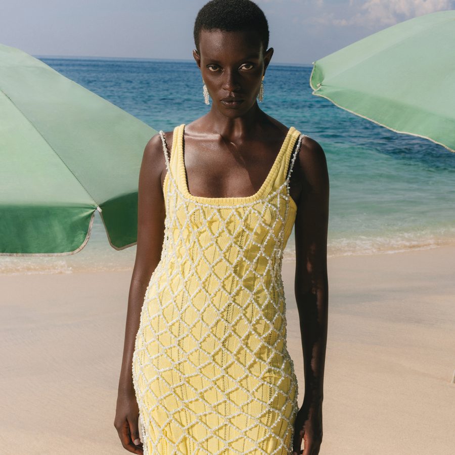 A woman stands by the beach in front of two beach umbrellas, wearing a yellow dress layered with a fishnet covering from Magali Pascal.