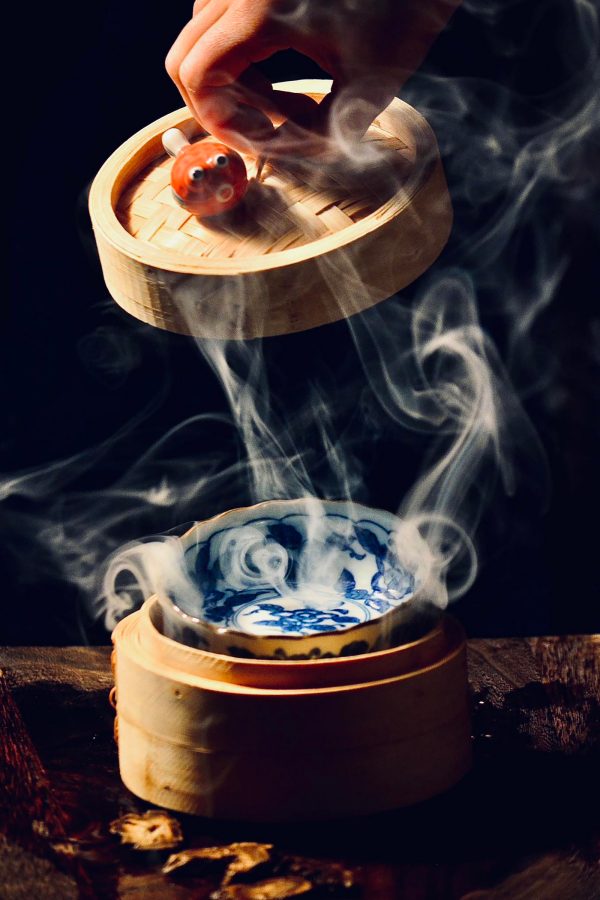  A moody shot of a hand lifting a bamboo steamer basket lid off a blue and white Chinese porcelain bowl with white mist, served at Healer in Shanghai.