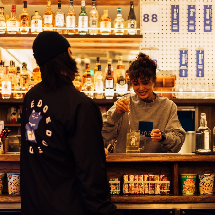 A person in black chatting with a smiling woman bartender while she mixes a drink at Lucky Mart, Shanghai.