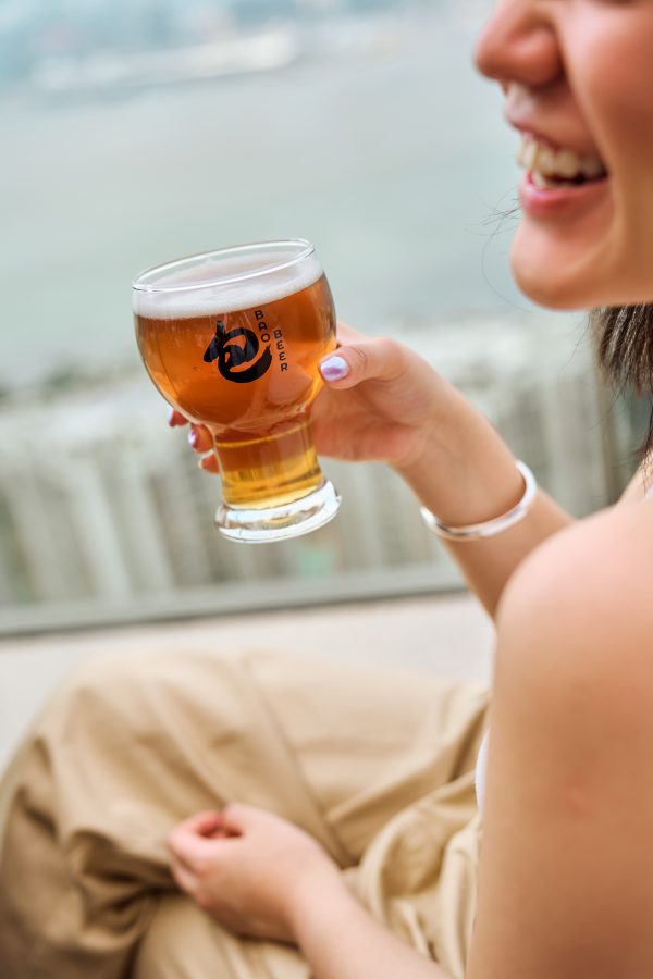 A woman smiles and raises a pint of Bao Beer at East Hong Kong with the harbour in the background.  