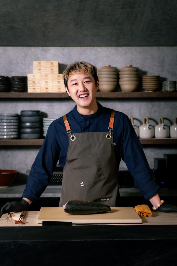 A chef smiles for the camera in a grey kitchen.  
