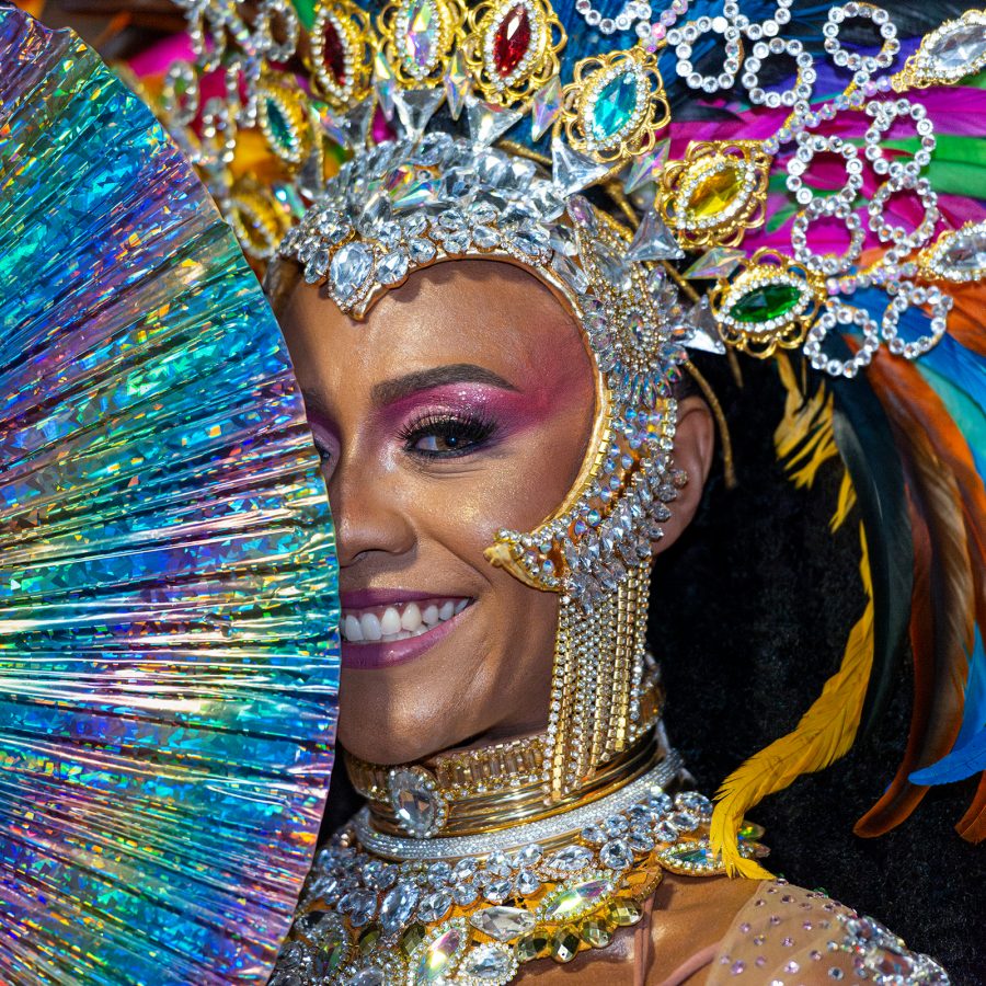 Woman dressed in rainbow carnival costume peeks out from behind a rainbow fan.