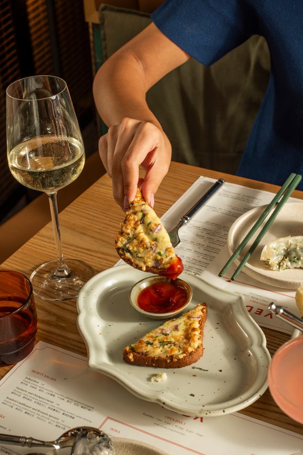 A hand holding a piece of bread over a plate at a dining table with dishes and a glass of white wine at Veda Cafe and bar.