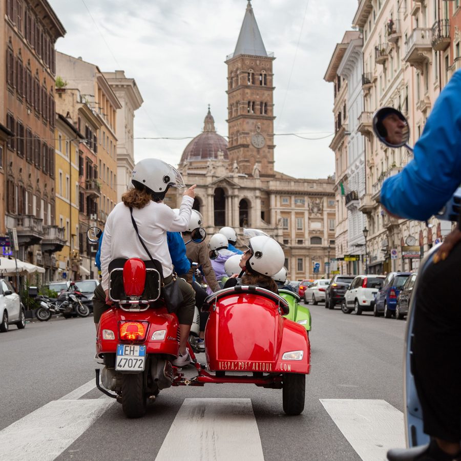 A red vespa with a side carriage drives through the streets of Rome.
