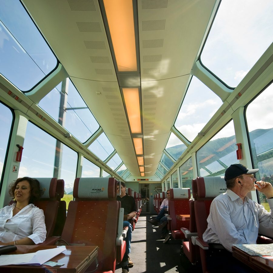 Two passengers sit on either side of a glass roofed train in Switzerland.  