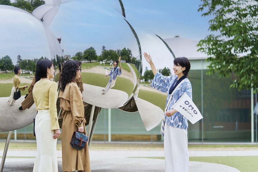 A woman working at OMO5 Kanazawa Katamachi by Hoshino Resorts holding a flag with the hotel logo and waving at two female guests. 