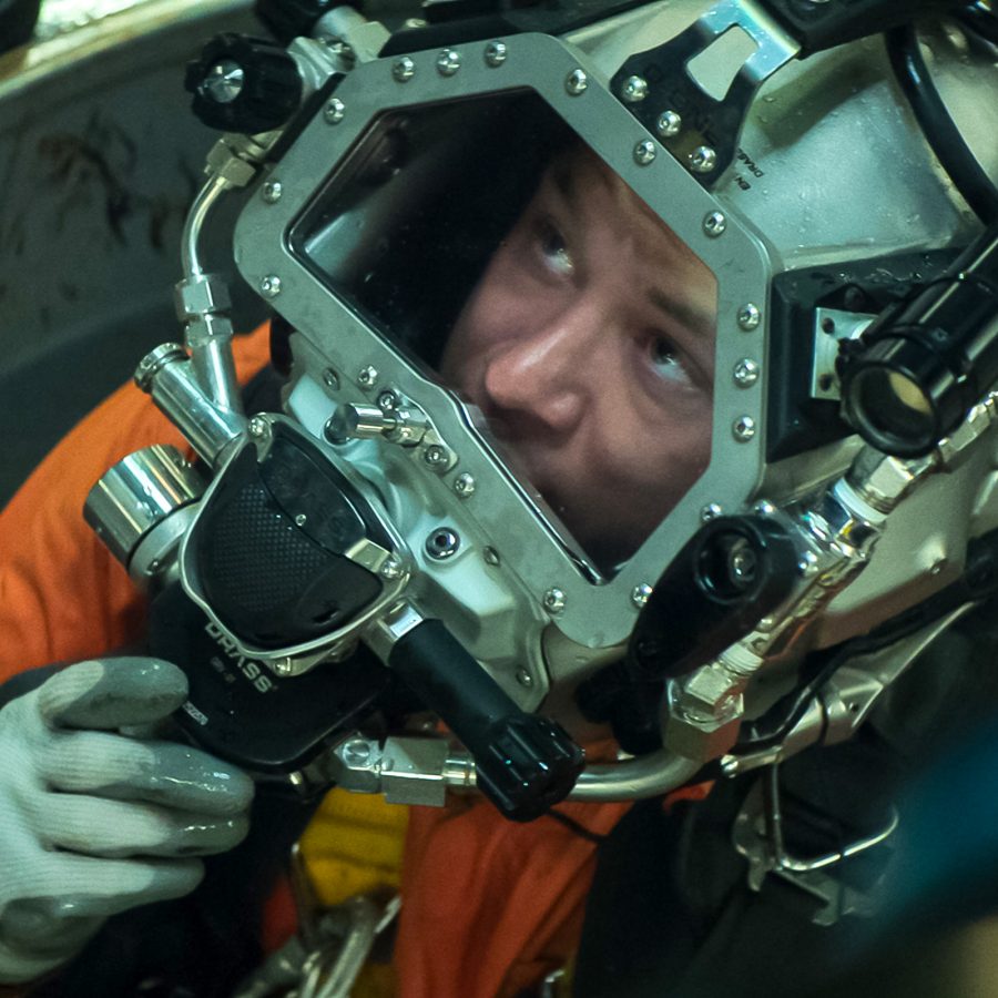 In a scene from Last Breath, diver Chris (Finn Cole) prepares to descend through a hole into the water. He wears an orange suit and a large breathing apparatus over his head. 