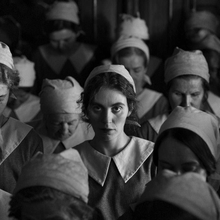 A woman dressed in a factory uniform looks up as she stands amid a crowd of other women looking down, in a black-and-white image from the film The Girl with the Needle.