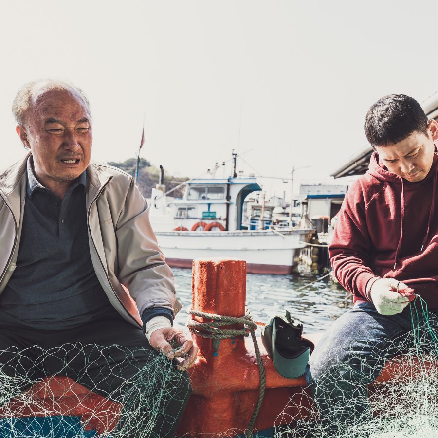 In a scene from The Land of Morning Calm, elder fisherman Yeong-guk (Yoon Joo-sang) and younger fisherman Yong-su (Park Jong-hwan) sit on the edge of a boat docked in a harbour; they are holding a large piece of net. 
