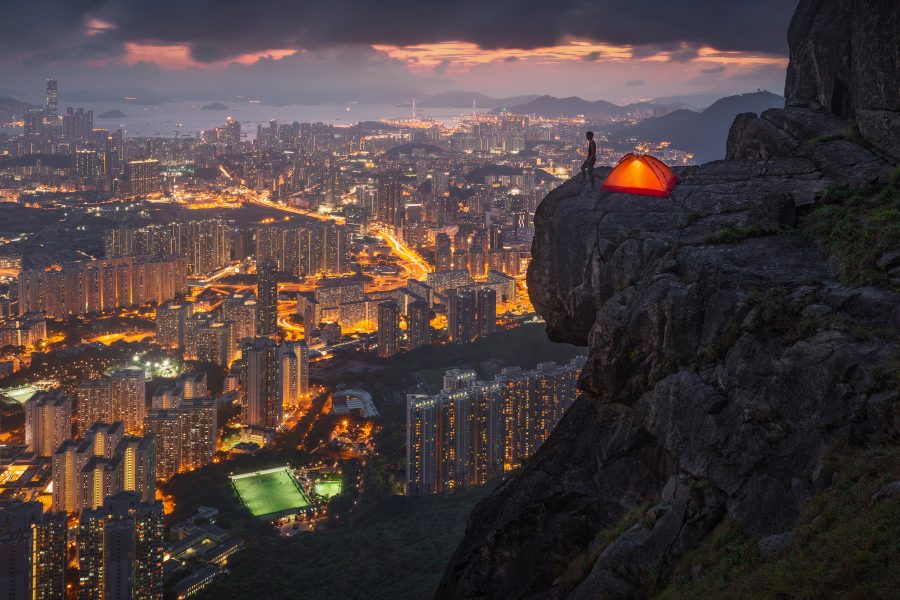 A person stands beside an illuminated tent on the edge of a cliff overlooking Hong Kong’s illuminated buildings below.