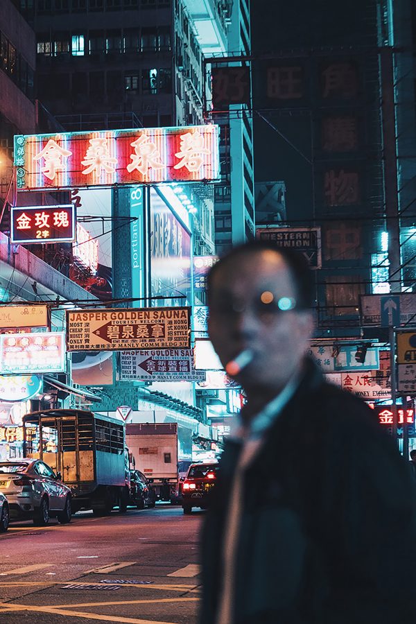 A man smoking a cigarette walks across the street against a street filled with neon in Hong Kong. 