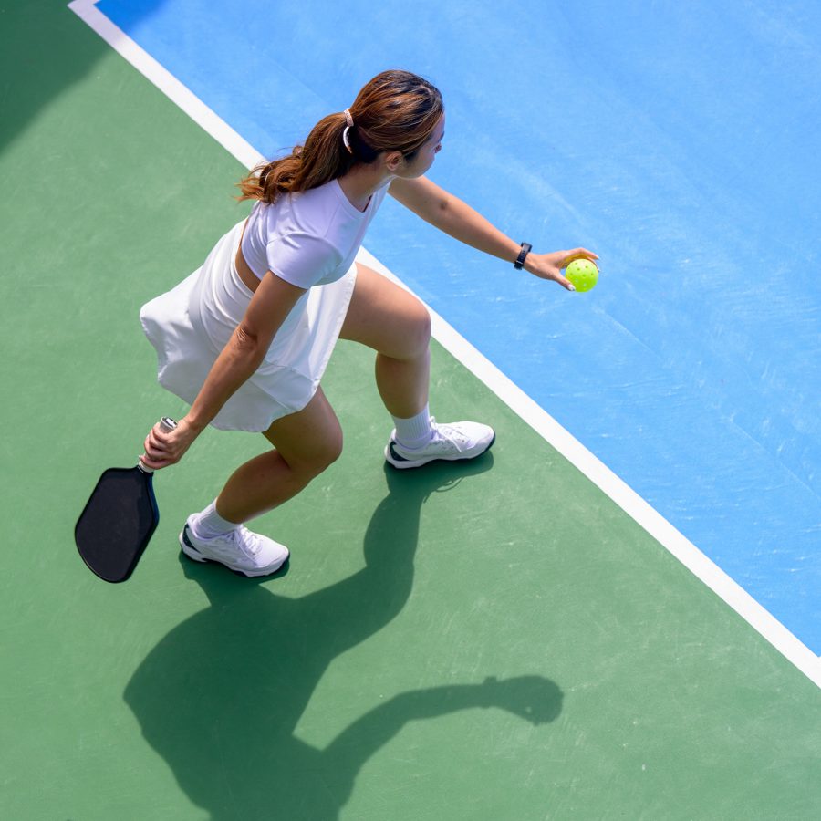 A female pickleball player gets ready to serve the ball at the baseline of the court. 