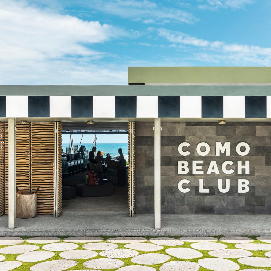 The exterior of Como Beach Club is a low-lying building with a striped roof, seen under a blue sky. 
