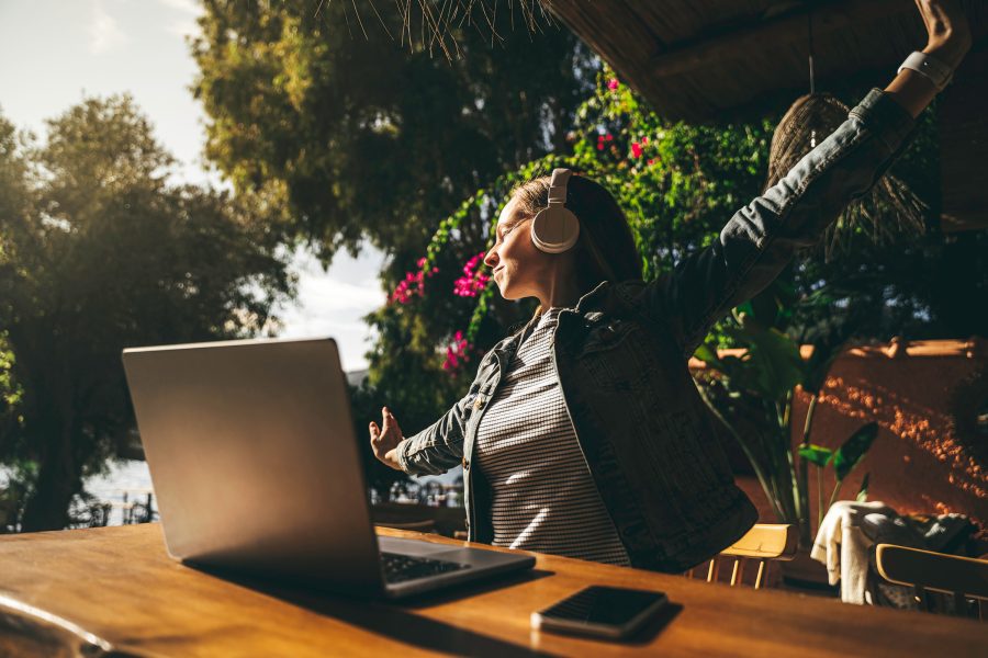 A remote worker with a pair of headphones on stretches as they work on a laptop outdoors.