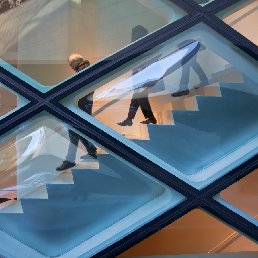 People visible through a diamond glass facade as they walk down a staircase.