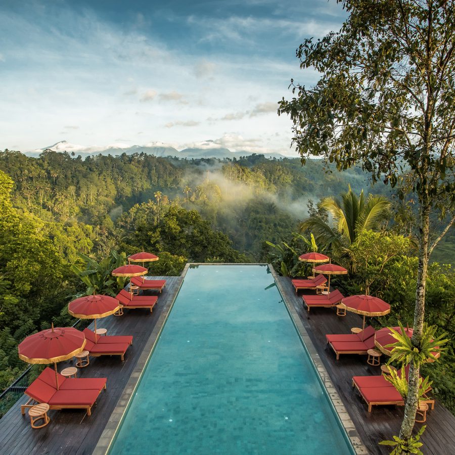An outdoor pool, sandwiched between two walkways decorated with red lounge chairs at Buahan, A Banyan Tree Escape, overlooks a lush jungle in Bali.