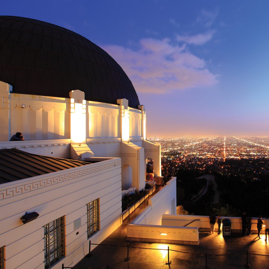 The white domed building of the Griffith Observatory is illuminated at night. In the distance, the lights of the city can be seen