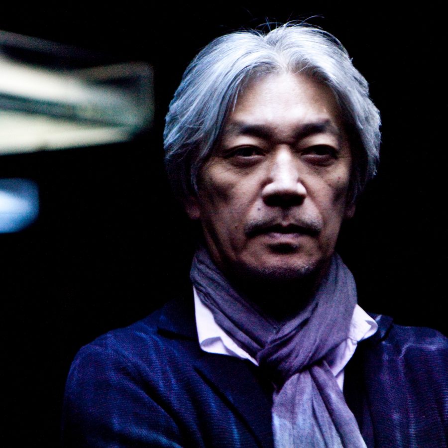 Ryuichi Sakamoto poses for a picture in front of a dark background. 