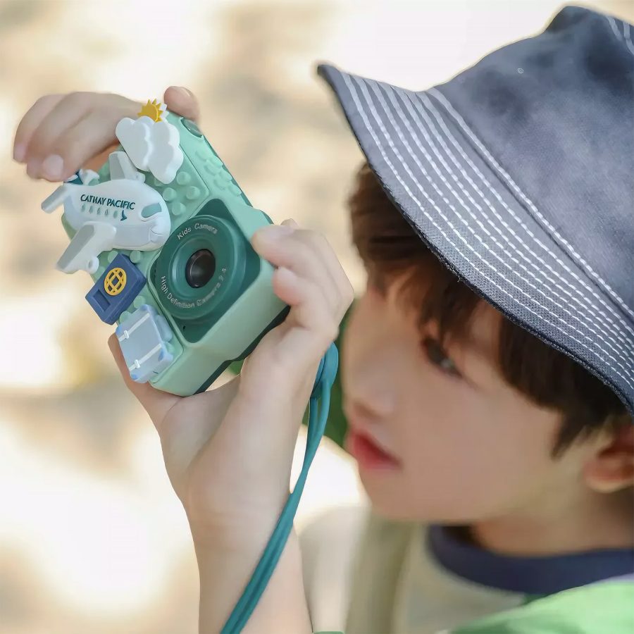 A young child wearing a blue bucket hat and holding a Cathay Pacific kid’s digital camera and pointing it upwards to take a photo 