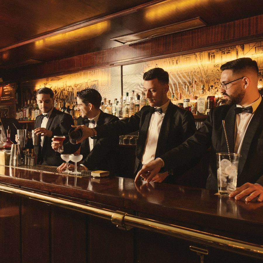 Interior view of five barmen in tuxedos making various cocktails behind a traditional wooden bar with a brass rail.