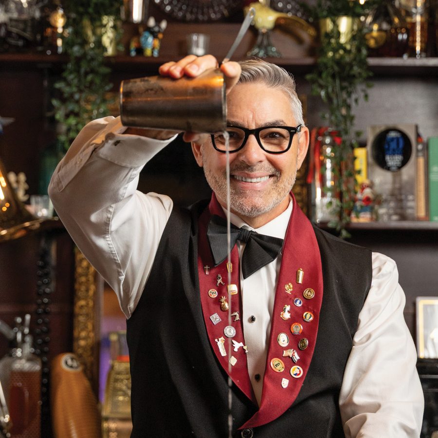 Portrait of a barman wearing a bowtie and a red-and-black waistcoat adorned with various pin badges pouring a cocktail out of a shaker
