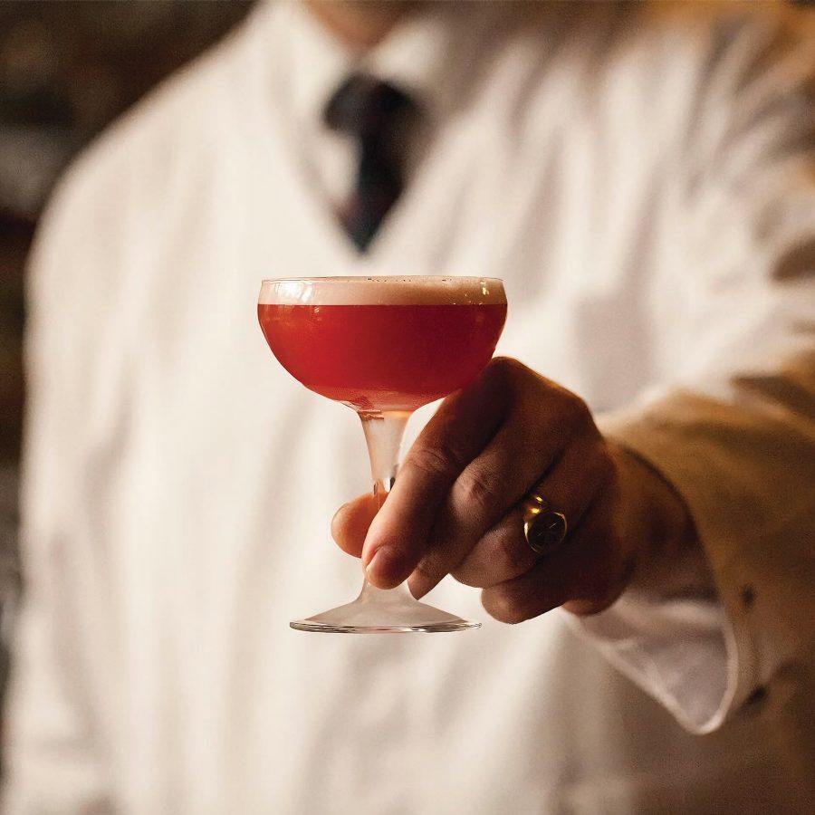 Close-up view of a bartender holding an orange-coloured cocktail in a short champagne glass.