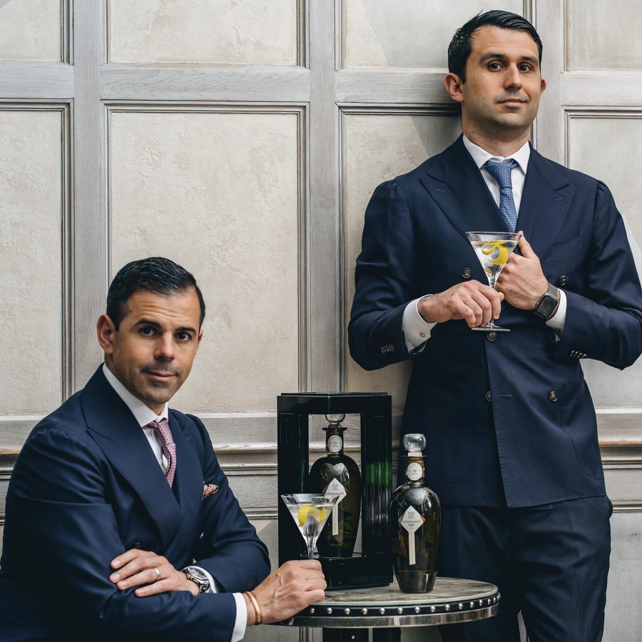 two Connaught mixologists – one sitting, one standing – in double-breasted blue suits holding martinis with lemon peel.